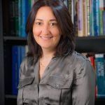 A woman is sitting in front of a bookshelf and smiling.
